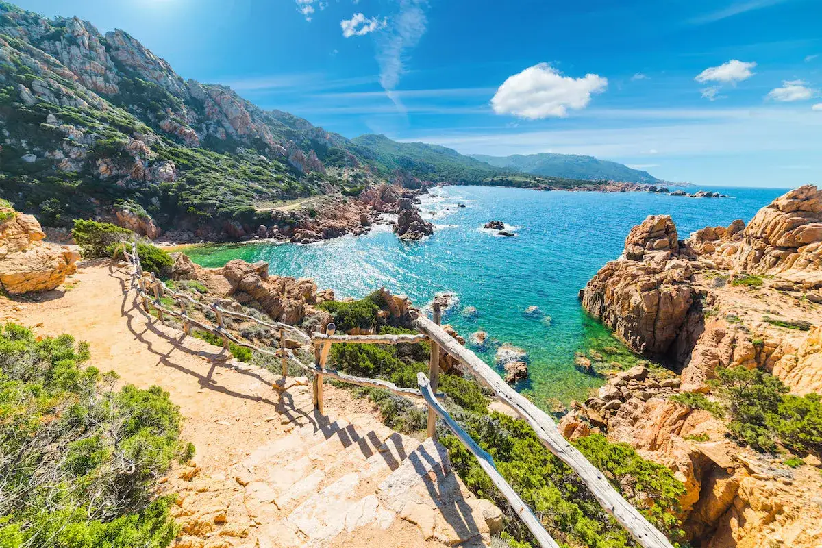 Sentier menant à une crique aux eaux turquoise, typique de la côte de Smeralda en Sardaigne. Rochers ocres et végétation luxuriante.