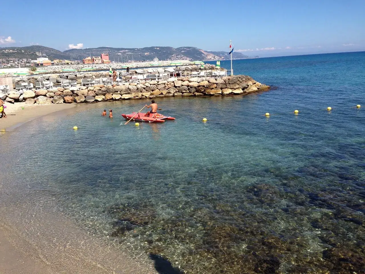 Un homme rame sur un kayak rouge à Loano, Italie, tandis que des enfants nagent dans les eaux claires.