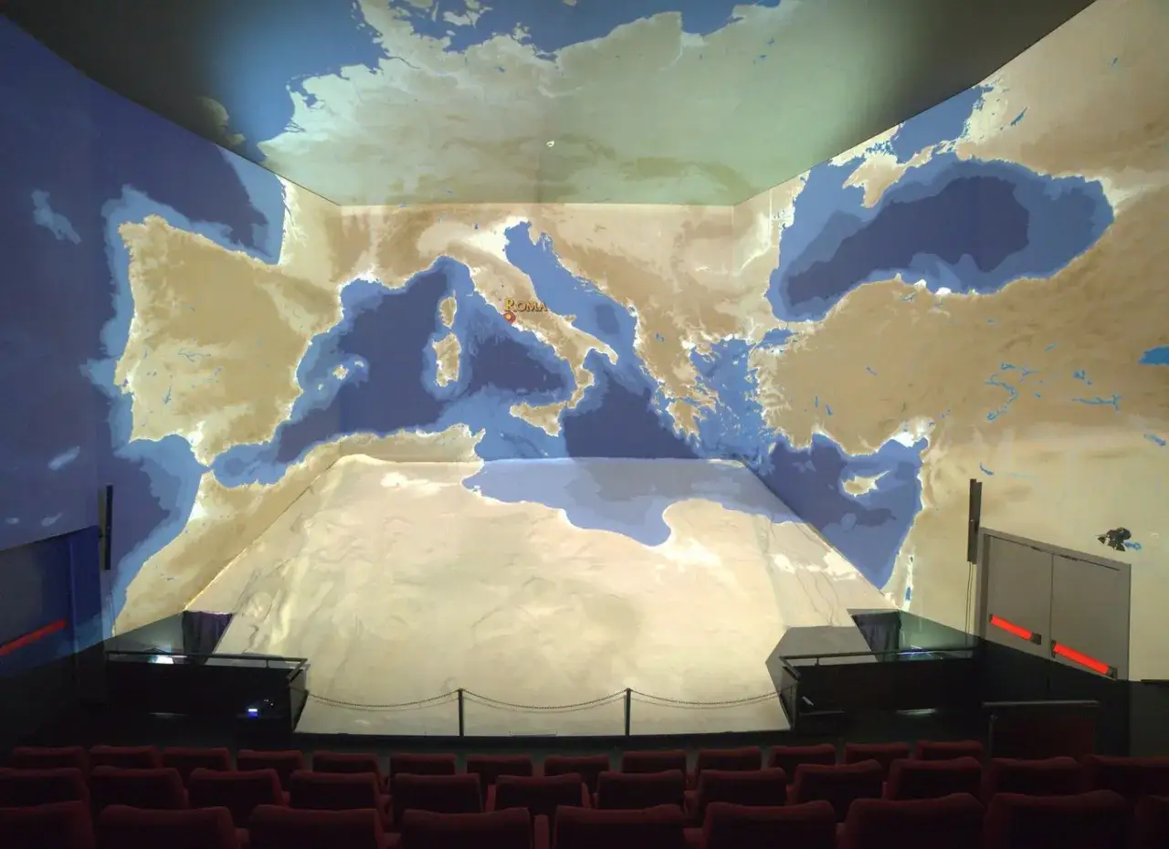 Salle immersive projetant une carte du monde antique, avec un point rouge marquant ROMA. L'ikono Rome est au centre de l'attention.