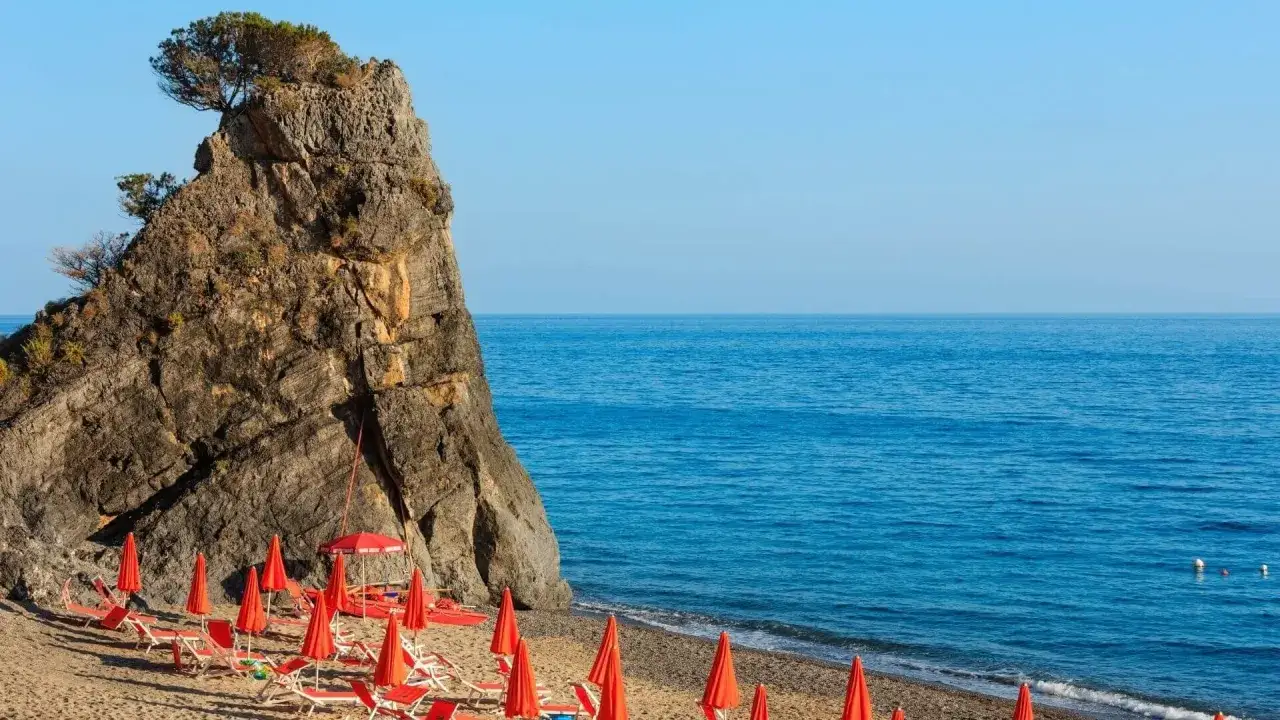 Plage d'Ascea Marina avec un rocher imposant, des chaises longues rouges et la mer bleue.