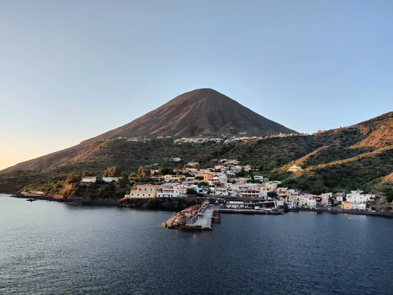Vue d'un village pittoresque sur l'&icirc;le Salina, avec un volcan imposant en arri&egrave;re-plan, baign&eacute; par la lumi&egrave;re dor&eacute;e du soleil couchant.
