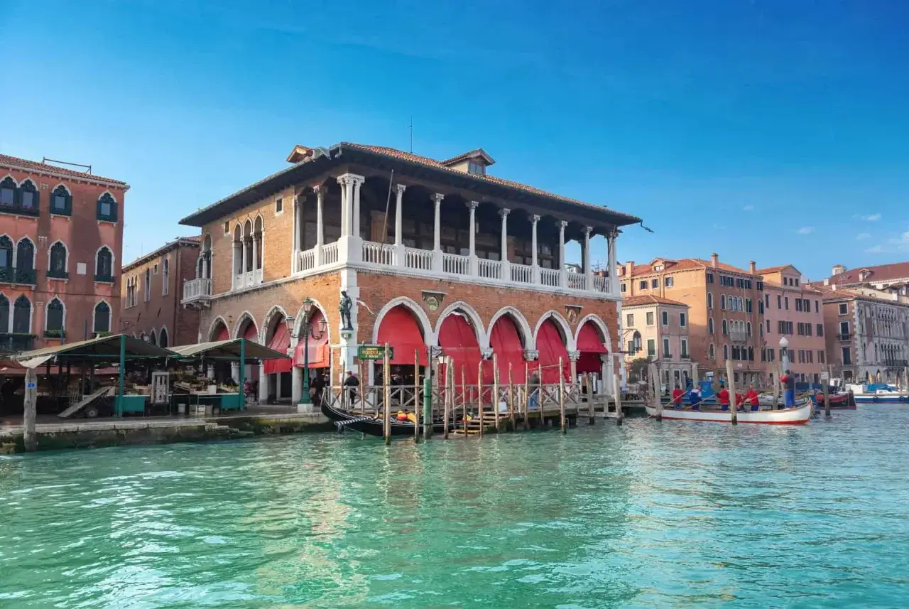Le marché du Rialto à Venise, avec ses arcades rouges et ses gondoles, s'anime sous un ciel bleu.