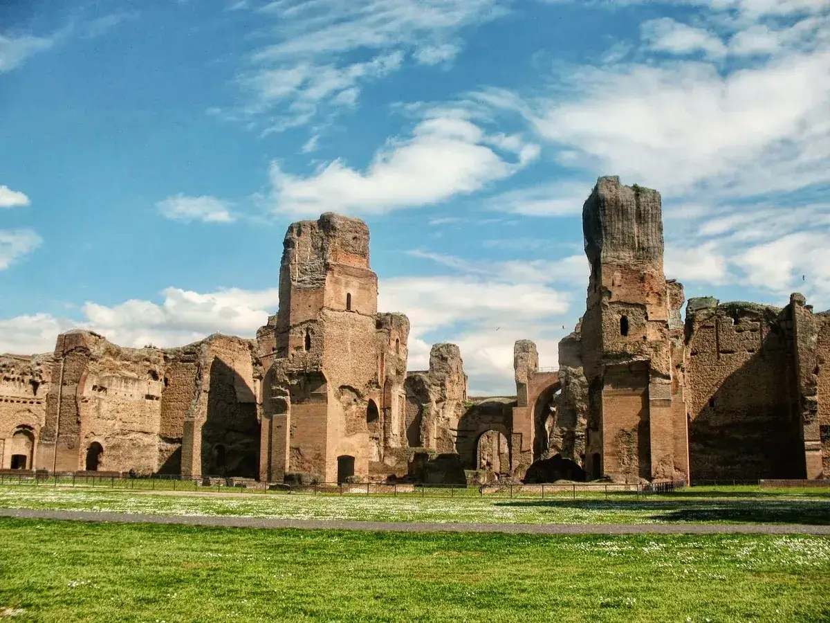 Ruines imposantes du bain de Caracalla sous un ciel bleu et nuageux. L'herbe verte et les petites fleurs bordent les vestiges antiques.