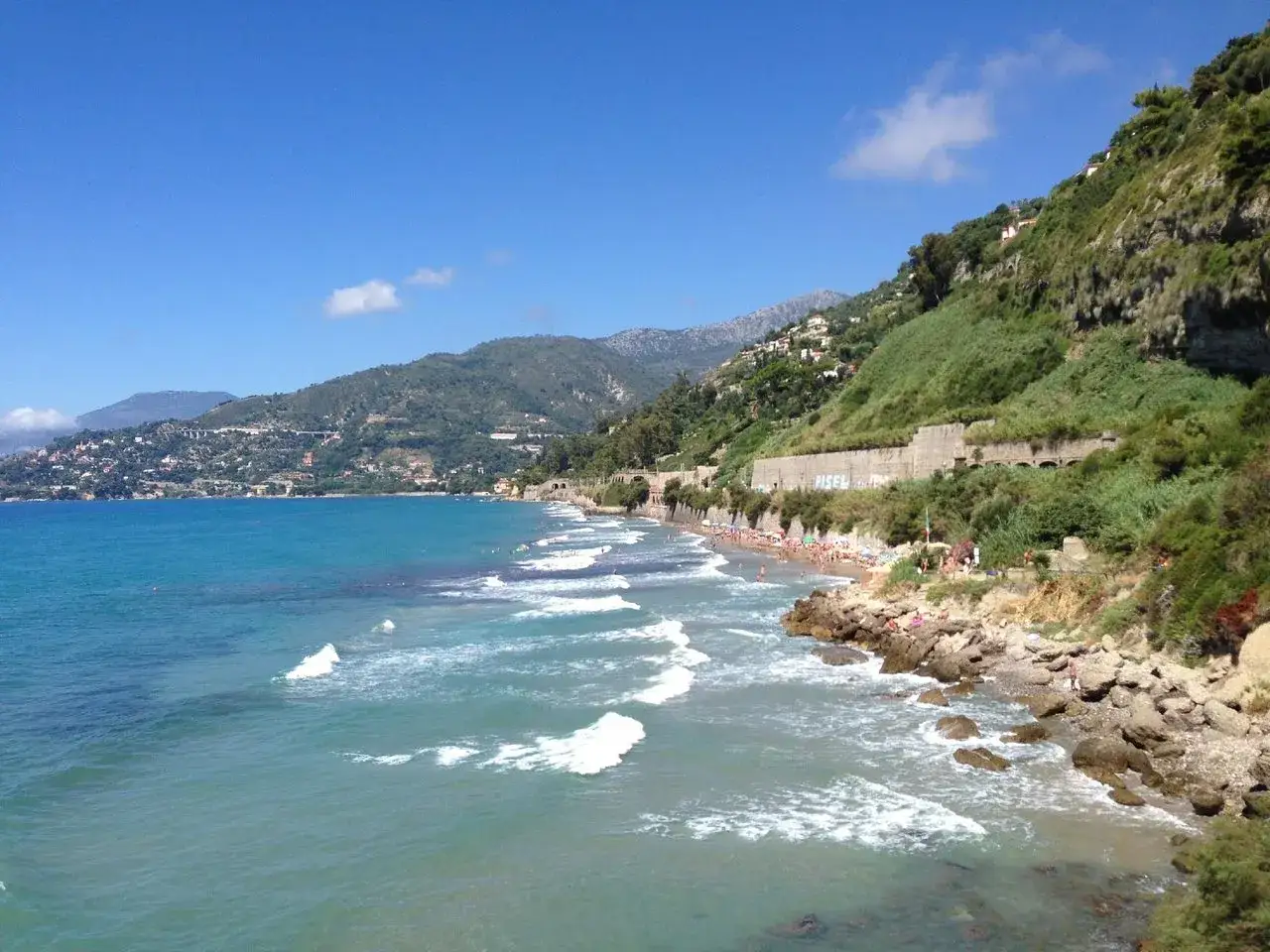 Plage animée à Ventimiglia, avec des vagues bleues et des collines verdoyantes en arrière-plan.
