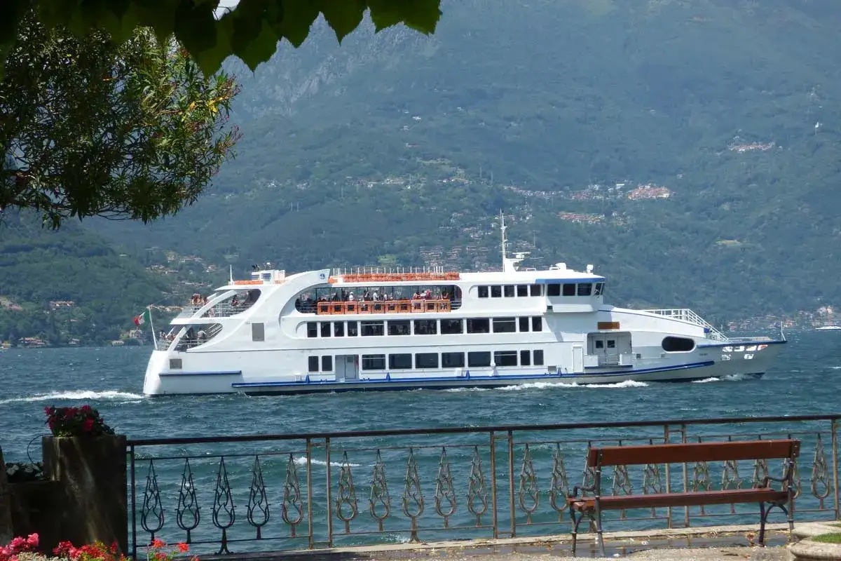 Un grand ferry blanc navigue sur le lac de Côme, transportant des passagers. Les montagnes verdoyantes forment un décor pittoresque.