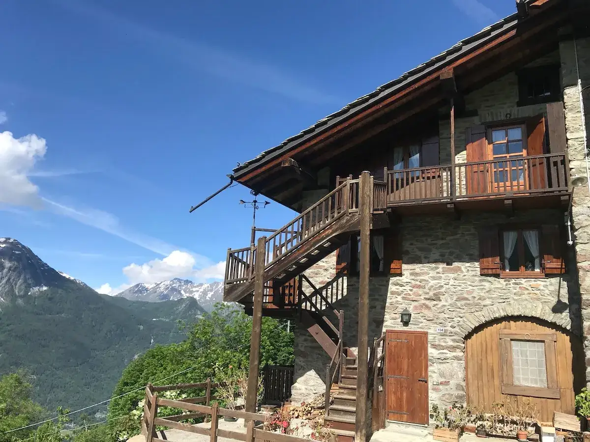 Maison de montagne en pierre avec escalier extérieur en bois, offrant une vue imprenable sur les Alpes. Idéal pour une location vacances Aoste, Italie.