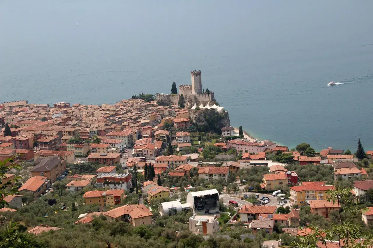 Vue panoramique de Malcesine, avec son château médiéval dominant le lac. Bateaux sur l'eau, maisons aux toits de tuiles rouges.
