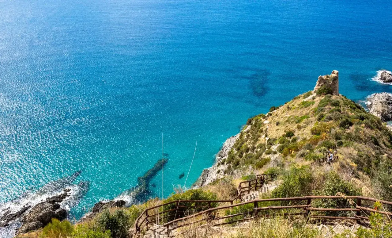 Vue panoramique d'Ascea Marina, avec la mer d'un bleu profond et une tour en ruine sur une colline verdoyante. Un sentier mène à la tour.