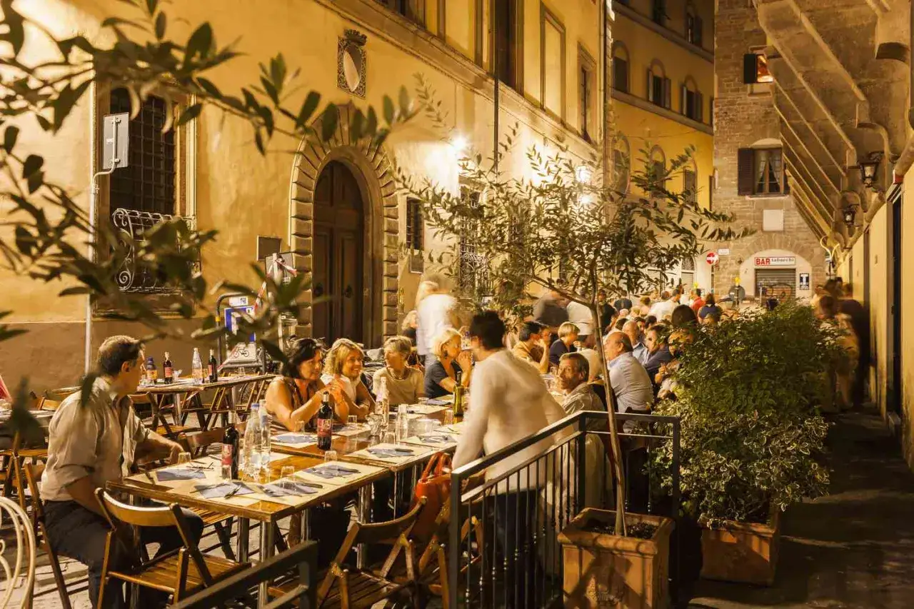 Terrasse de restaurant animée à Florence le soir, avec des gens attablés sous des arbres.