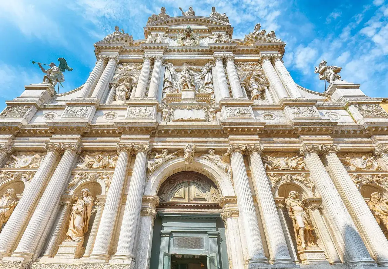 Façade baroque de Santa Maria del Giglio à Venise, ornée de statues et de colonnes imposantes, sous un ciel bleu éclatant.