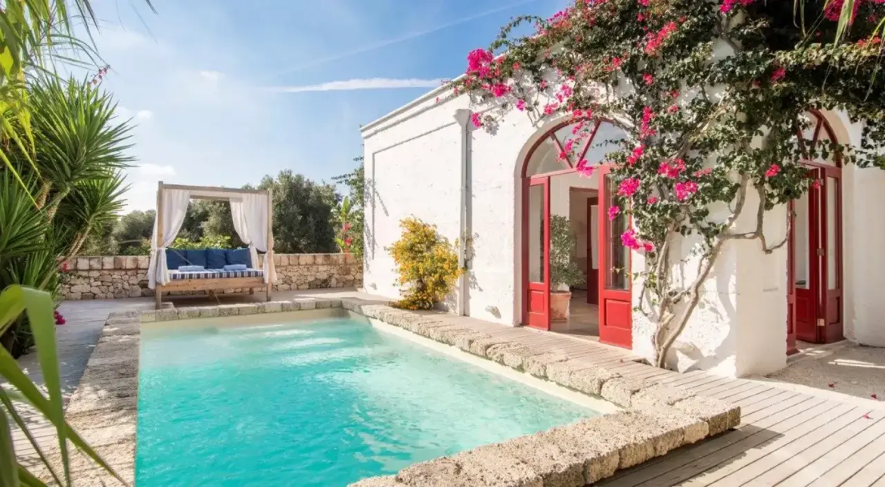 Une piscine turquoise devant une masseria en Italie, avec des bougainvilliers roses grimpant sur le mur blanc et une banquette de repos.