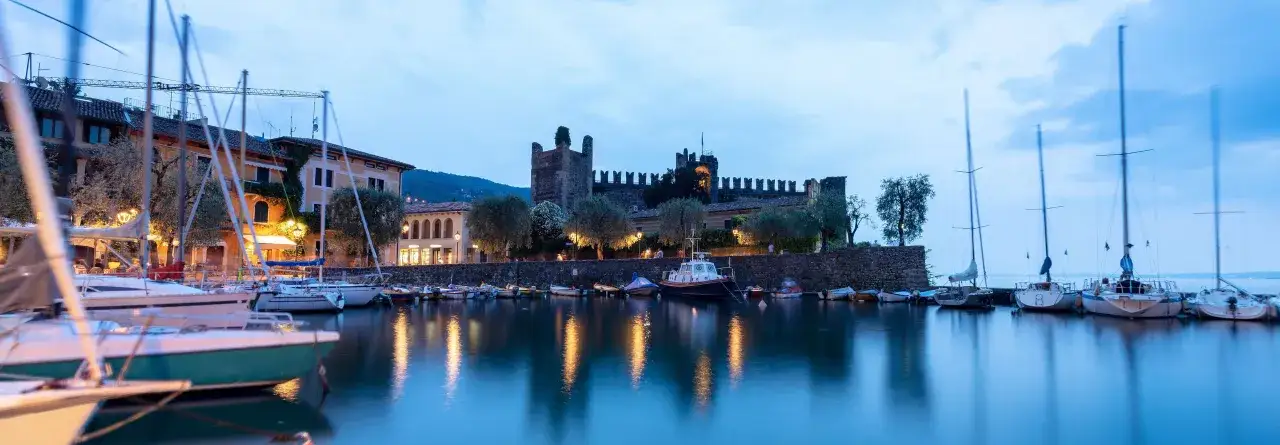 Port de plaisance de Torri del Benaco au crépuscule, avec des voiliers amarrés et un château médiéval illuminé en arrière-plan.