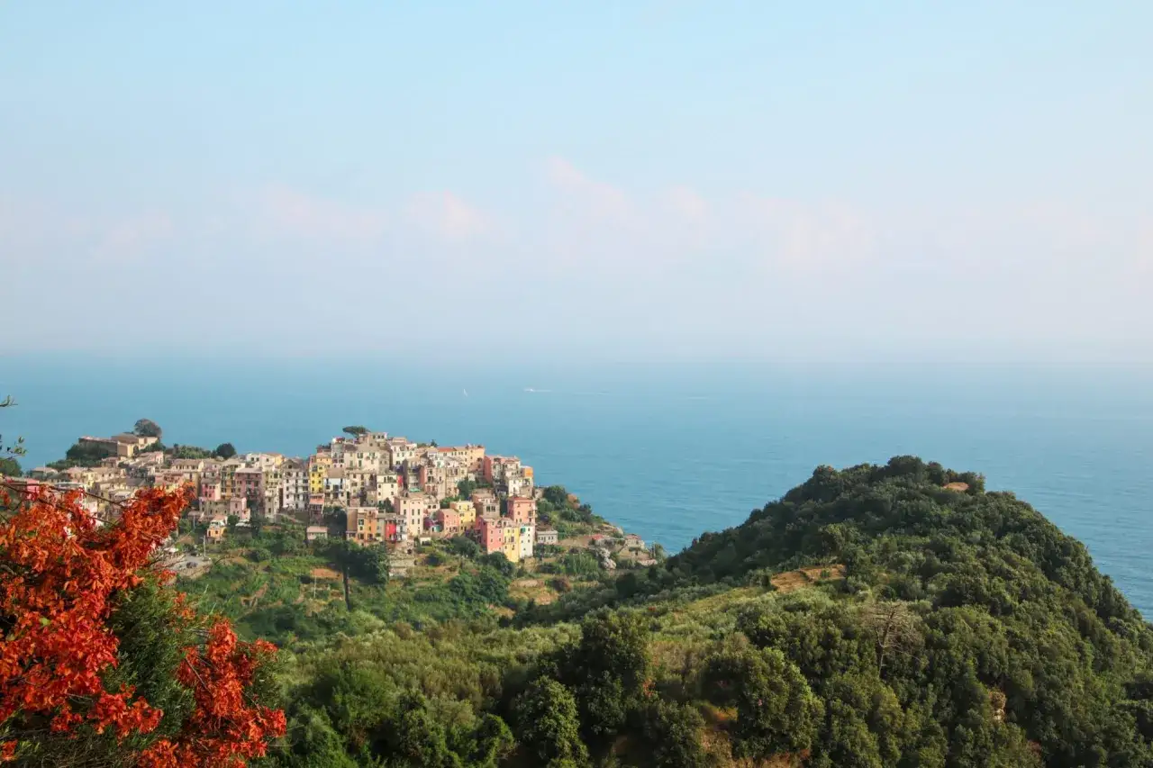 Vue panoramique de Punta Corvo, un village coloré accroché à flanc de colline, surplombant la mer bleue. Des arbres verdoyants et des feuilles orangées encadrent la scène.