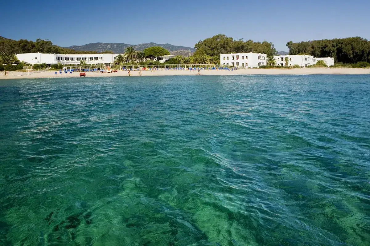 Eaux turquoise cristallines devant la plage de Pula en Sardaigne. Des hôtels blancs bordent le sable, avec des parasols et des gens profitant du soleil.