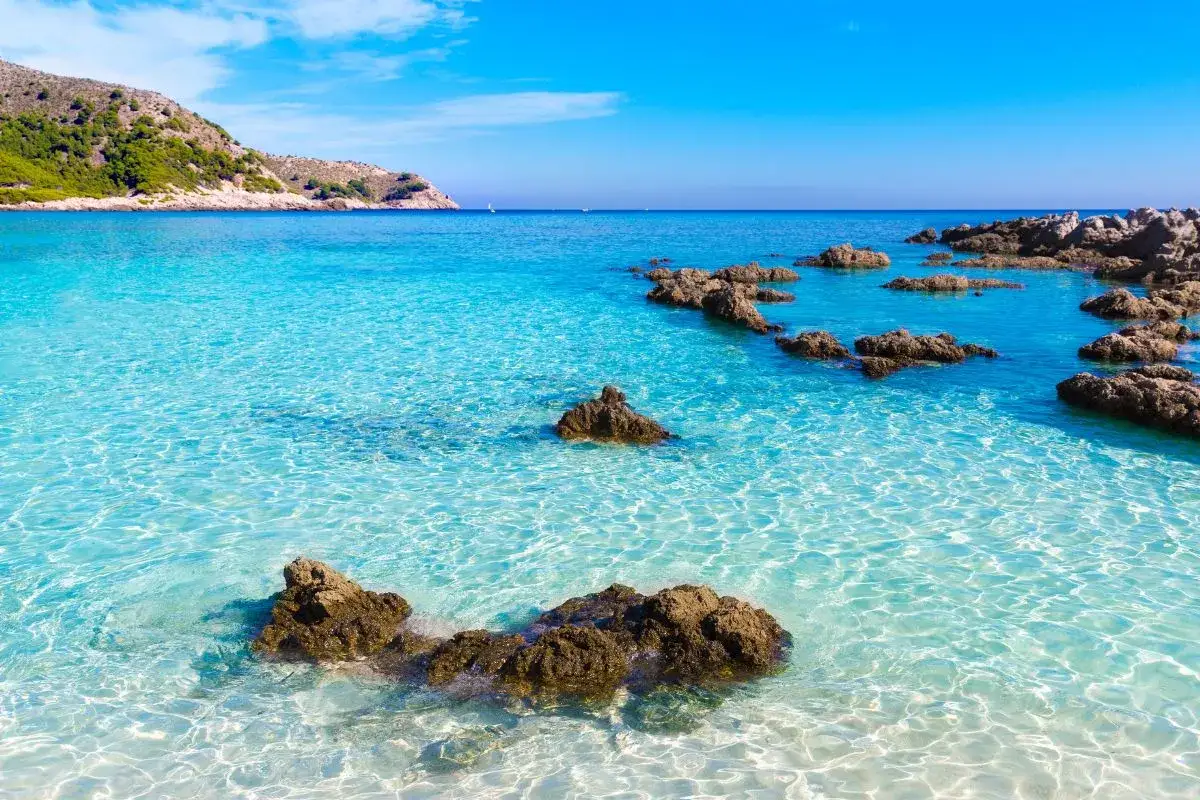 Eau turquoise cristalline, rochers sombres émergeant de la mer, colline verdoyante en arrière-plan. Un rêve de toscane plage.