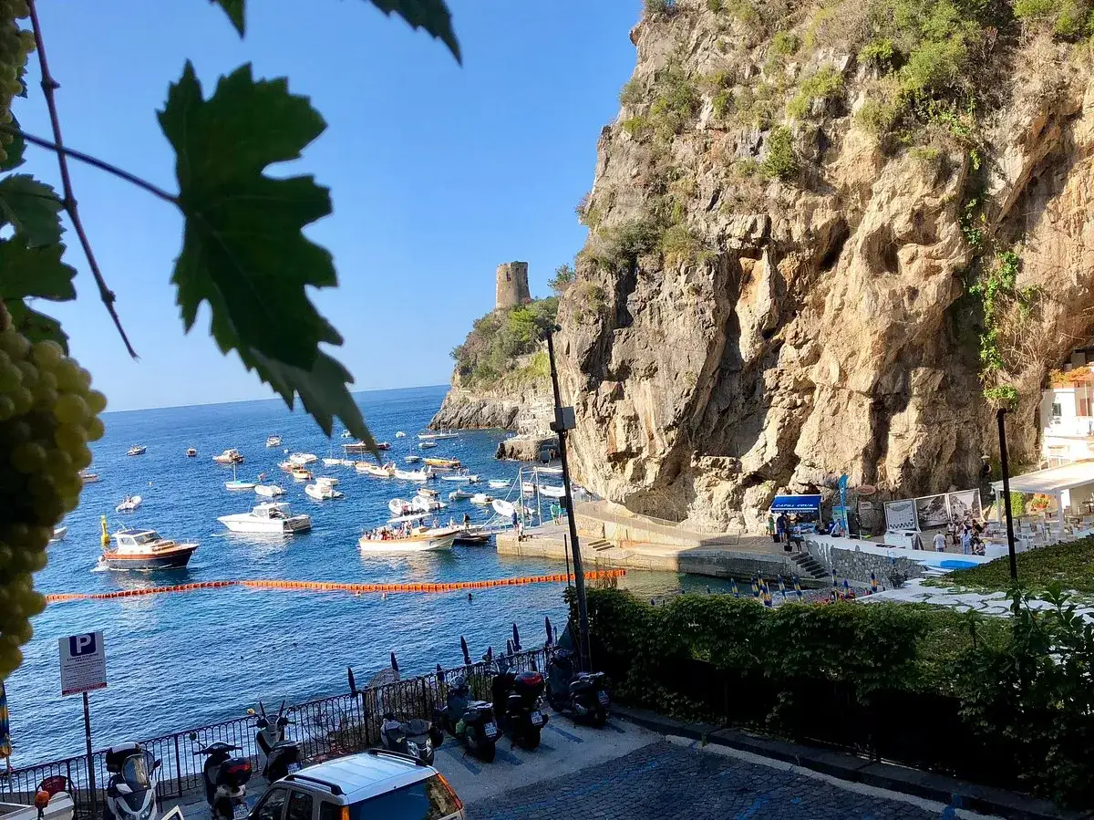 Vue pittoresque de Praiano, Italie, avec des bateaux dans la baie, une tour médiévale perchée sur la falaise et des vignes au premier plan.