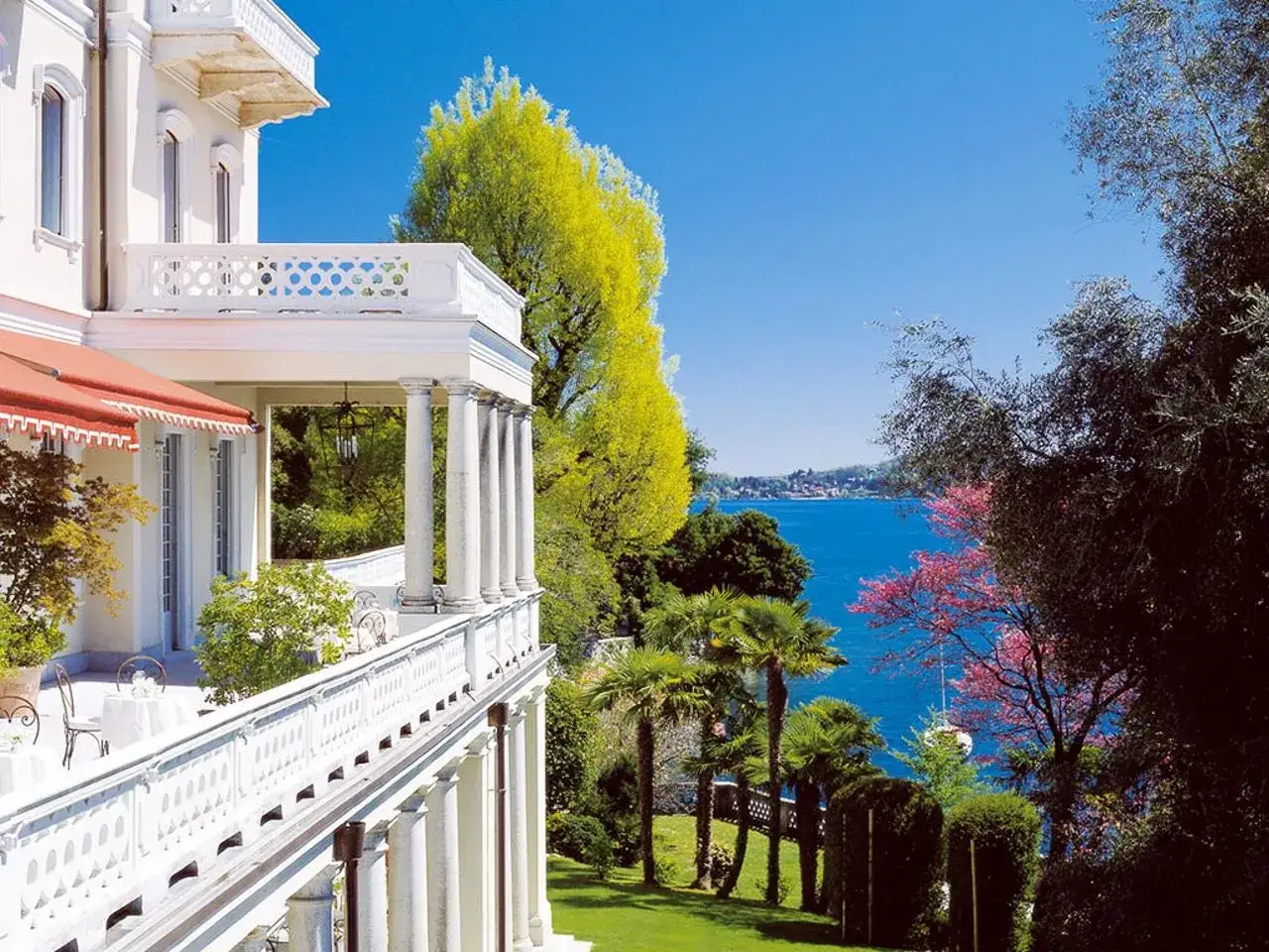 Magnifique h&eacute;bergement lac majeur avec balcon blanc, vue sur le lac bleu et v&eacute;g&eacute;tation luxuriante.