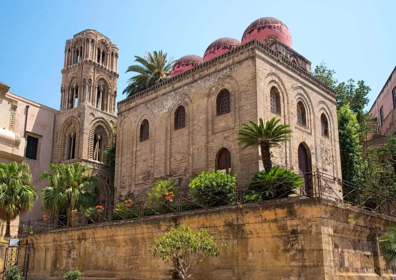 La capitale de la Sicile, Palerme, dévoile son église byzantine aux dômes roses et son campanile orné, sous un ciel bleu éclatant.