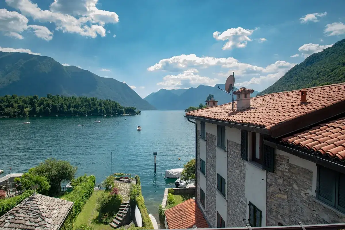 Vue d'une chambre d'hôte en Lombardie, avec un lac paisible, des montagnes verdoyantes et un ciel bleu parsemé de nuages.
