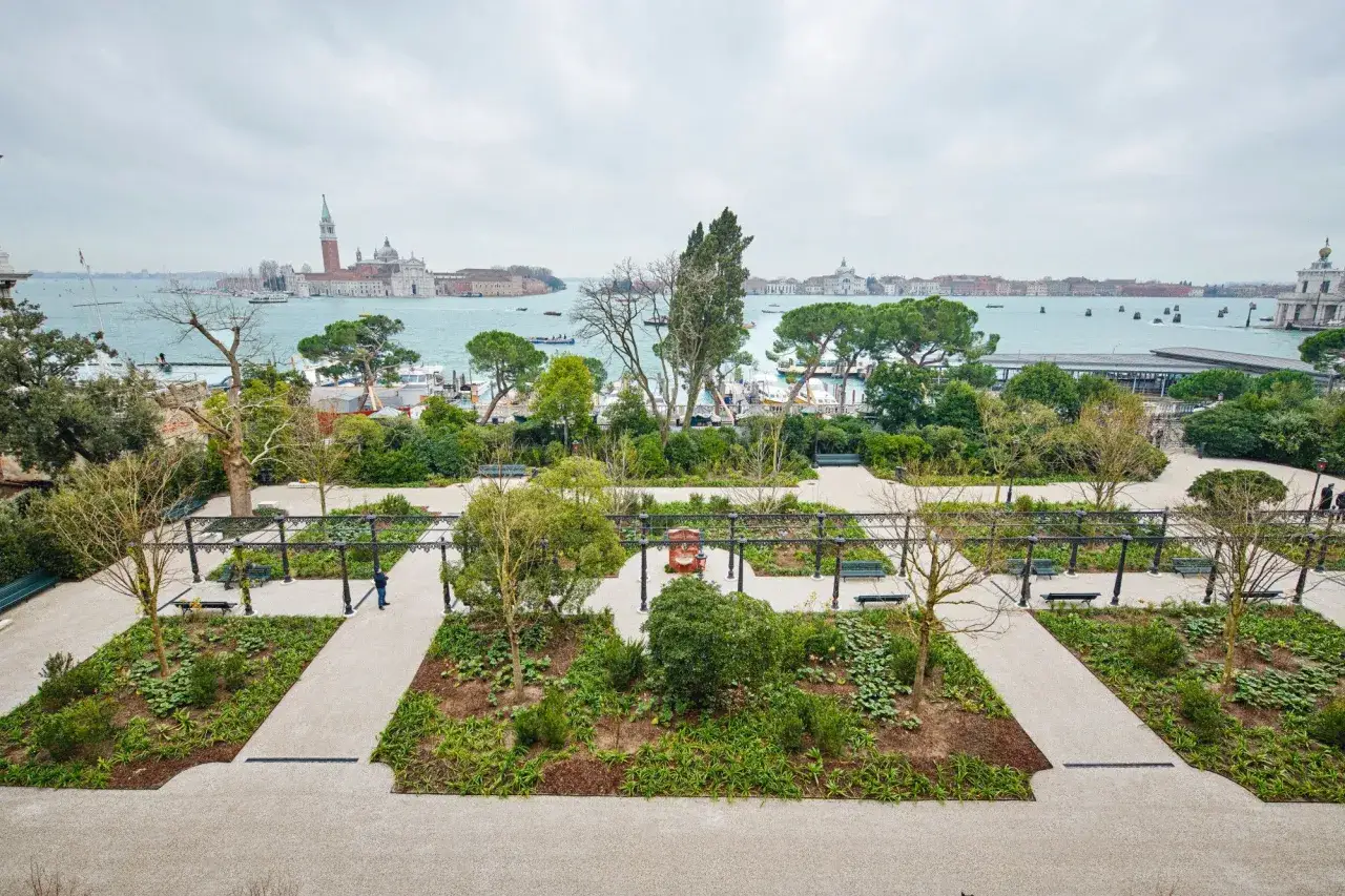 Vue d'un jardin à Venise, avec des parterres de fleurs, des pergolas et des bancs, donnant sur le lagon et les bâtiments historiques.