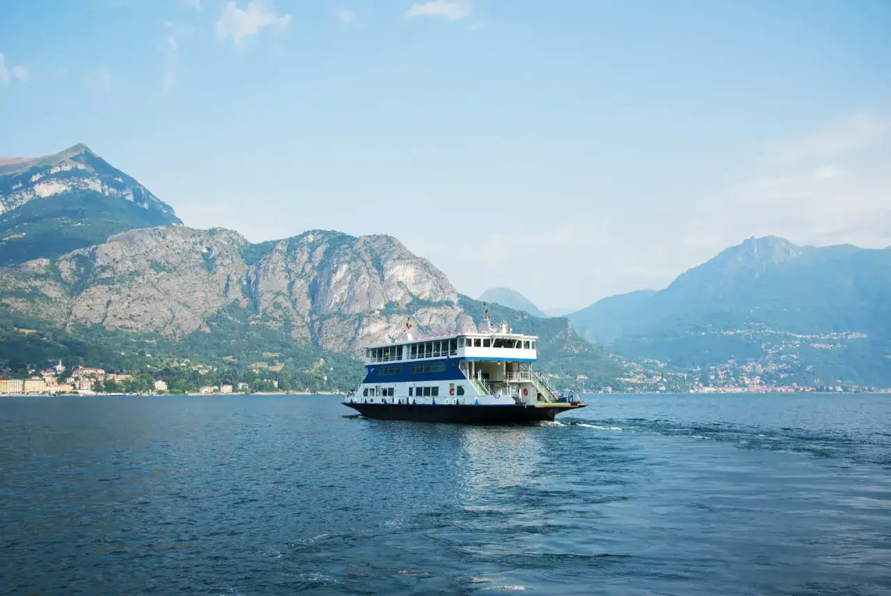 Un ferry transporte des voitures sur le lac de Côme, naviguant paisiblement entre des montagnes majestueuses sous un ciel bleu.