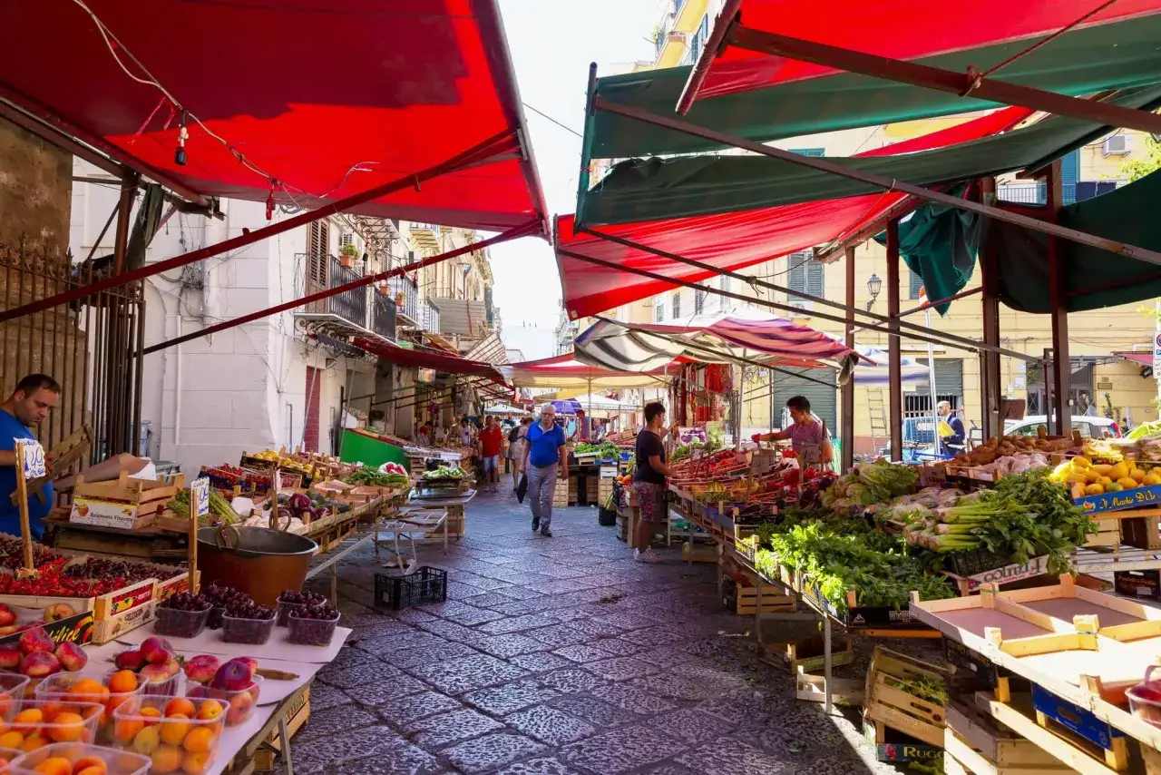 Ambiance animée d'un jour de marché à Taormina, avec des étals colorés de fruits et légumes frais sous des auvents rouges et verts.