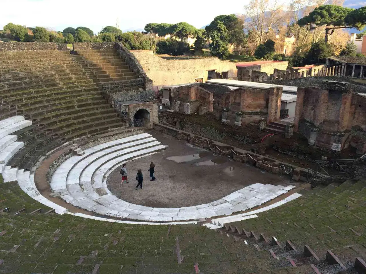 Trois personnes explorent une ancienne arène en Italie, ses gradins de pierre et ses ruines témoignant de son histoire.