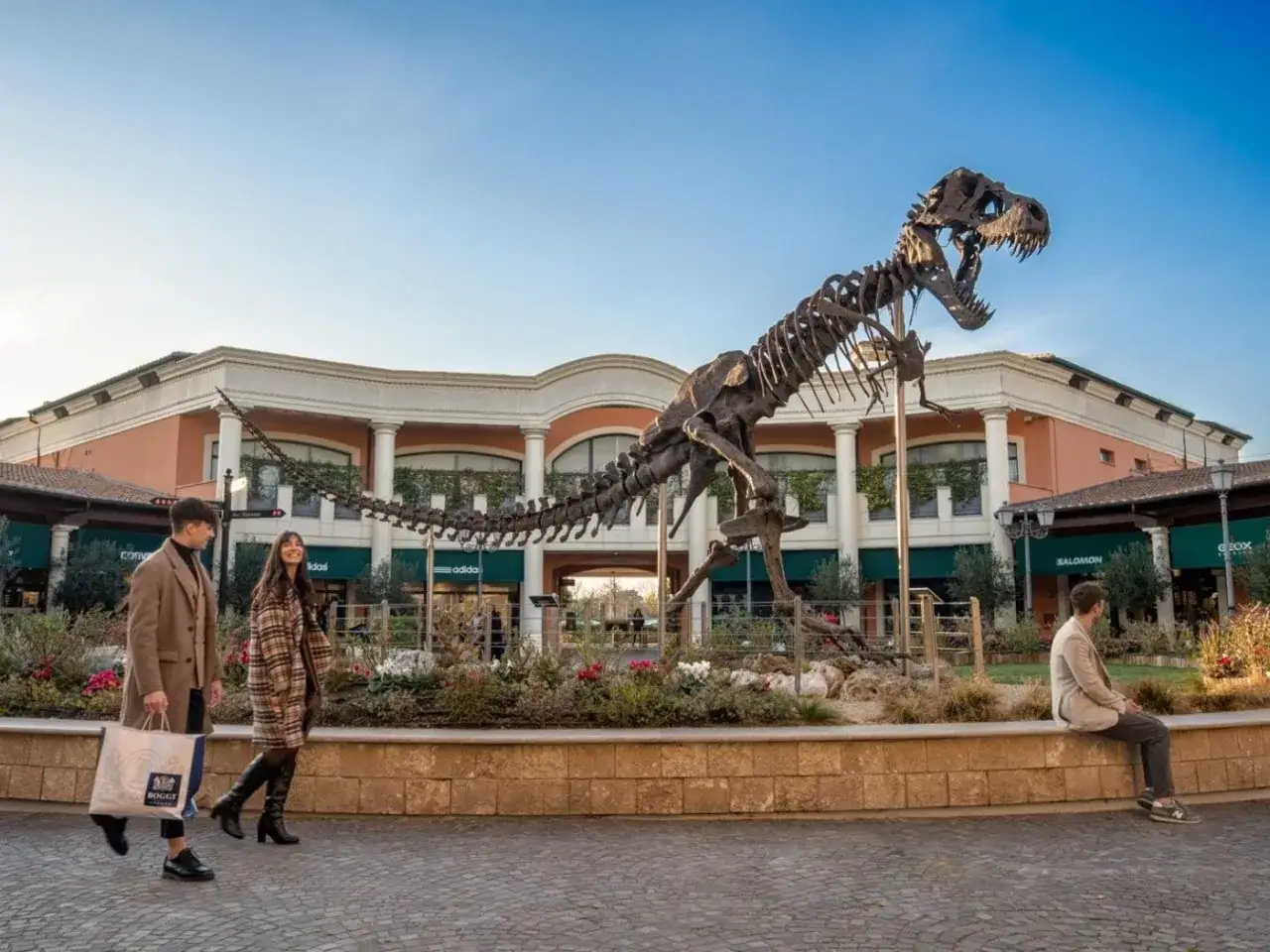 Un couple se promène devant un squelette de dinosaure au Castel Romano Outlet, avec des boutiques en arrière-plan.